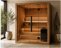 A wooden indoor sauna with glass doors, built-in wooden steps, warm ambient lighting, and a black sauna stove on the right side, shown in a styled bathroom setting with a plant, towel, and basket.