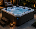 An outdoor rectangular hot tub with dark wood-look siding and a light top rim, filled with bubbling blue water and lit by built-in and ambient LED lighting. It is photographed on a patio at night with steps, a lantern, plants, and a patio sofa in the background.